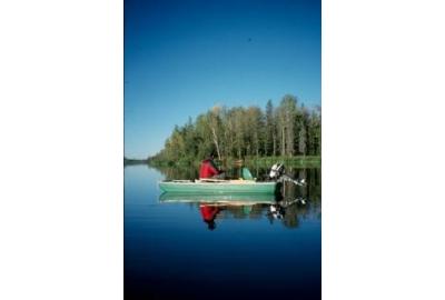 small green boat with man sitting and fishing on blue lake trees in the distance