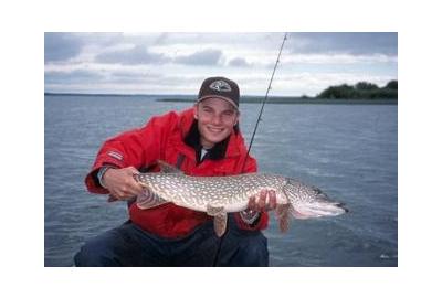 boy wearing hat and red jacket smiling holding a pike sitting by the water with fishing rod under his arm