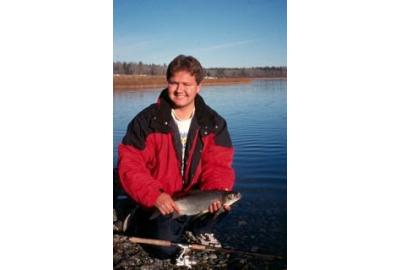 man in red jacket sitting on rocky shore holding fish with both hands