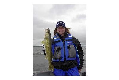 person wearing blue life fishing vest stands in boat holds a green bass fish with her right hand on a cloudy day