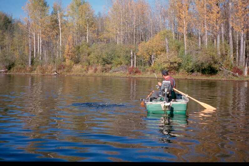 man in small green boat with oars in lake with summer to fall transition treeline