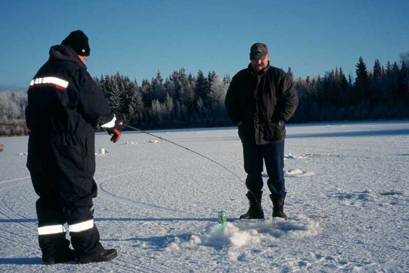 2 men standing by ice fishing hole one has casted their rod in