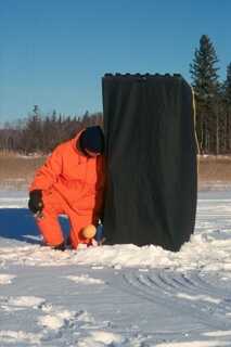 black ice fishing tent on snow man in orange snowsuit kneeling on its right side