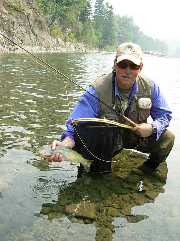 man squatting in shallow water holding fish in right hand and fishing rod in left