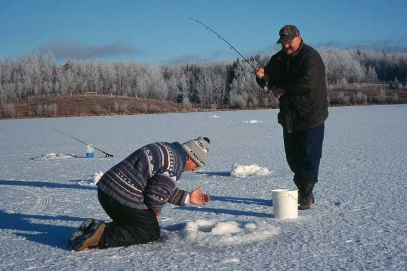 man kneels over to look into ice fishing hole adjusting line while another man is standing holding a fishing rod 