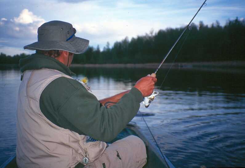 man in fishing vest and hat facing away from camera and fishing sitting on boat