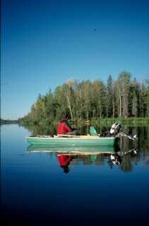 small green boat with man sitting and fishing on blue lake trees in the distance