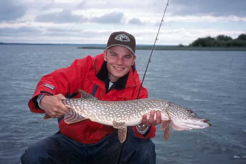 boy wearing hat and red jacket smiling holding a pike sitting by the water with fishing rod under his arm