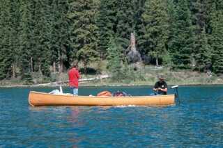 two men on opposite ends of a long boat in lake with coniferous forest in the background