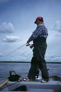 man standing on edge of boat fishing with rod pointing down towards the water