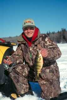 man wearing camouflage snow suit smiles and kneels down on snow with fishing rod in one hand and small fish on the other