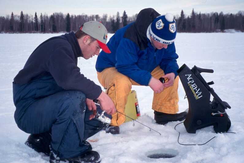 two men ice fishing one is holding a fishing rod and the other is operating an aqua vu machine