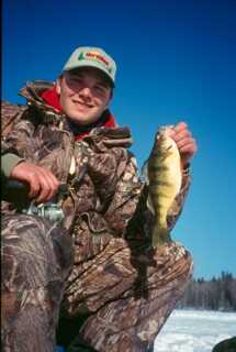 man wearing camouflage winter suit holds a small perch by its mouth 