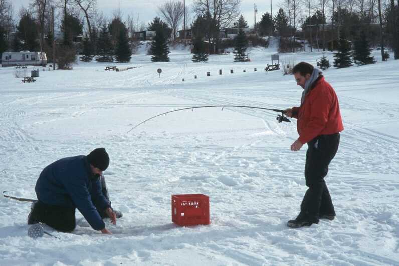 frozen river with one man examining ice fishing hole next to a red milk crate and another man is holding a long fishing rod to the left