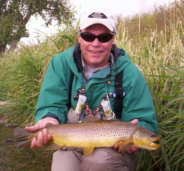 man wearing sunglasses holds a brown trout sitting by tall grass and dirt trail