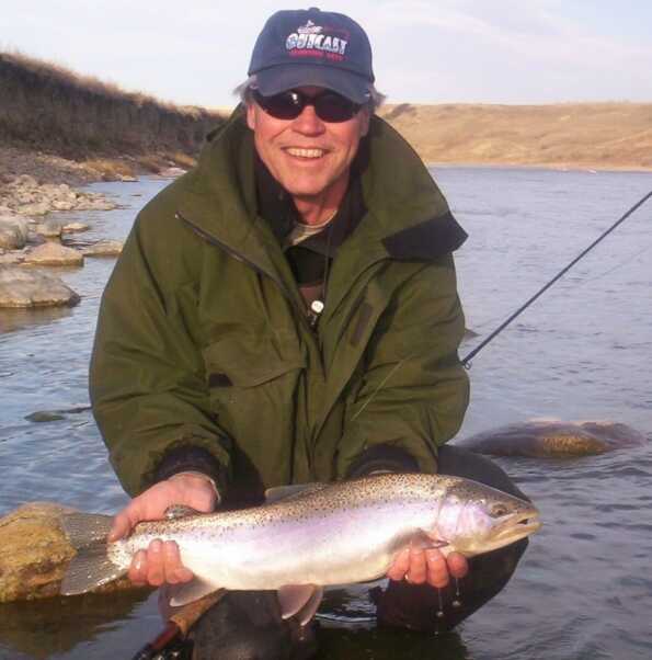 man wearing sunglasses and dark green jacket squats in a stream while holding a rainbow trout