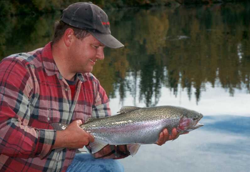 sideview of man holding large rainbow trout with both hands and lake reflecting sky is in the background