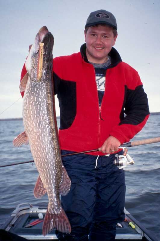 standing man wearing red jacket holds a large pike vertically with right hand and fishing rod in left hand