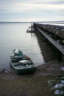 vertical shot of small fishing boat docked on shore next to a large wooden dock sky is cloudy and nearly sunset