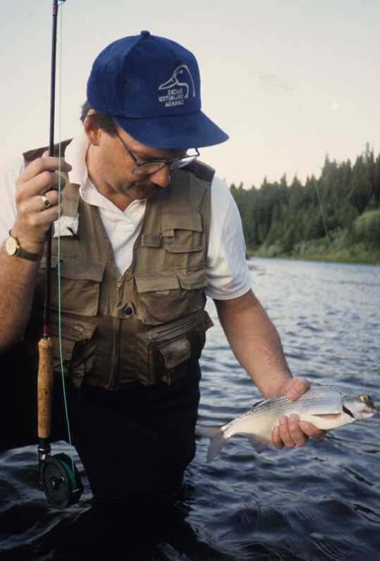 man standing in water holds fishing rod in right hand and grips on to small fish with left hand