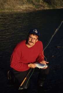 guy kneeling on one knee holding small fish in palm on hand and fishing pole leaning on his left arm