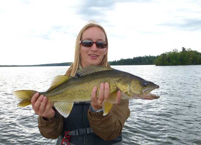 walleye fish held by blonde woman wearing sunglasses standing in front of water