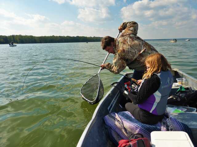 two sitting in a fishing boat girl is holding a fishing rod towards the water and man is holding a large net into the water